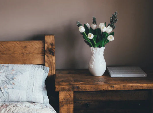 Picture of the corner of a bedroom with an unmade bed with grey bedding on it and a nightstand sitting next to the bed with a lamp and coffee mug on it