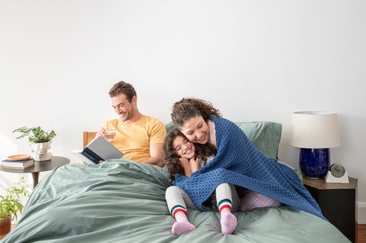 Family of three cuddling, reading and laughing in a bed made with sage colored bedding