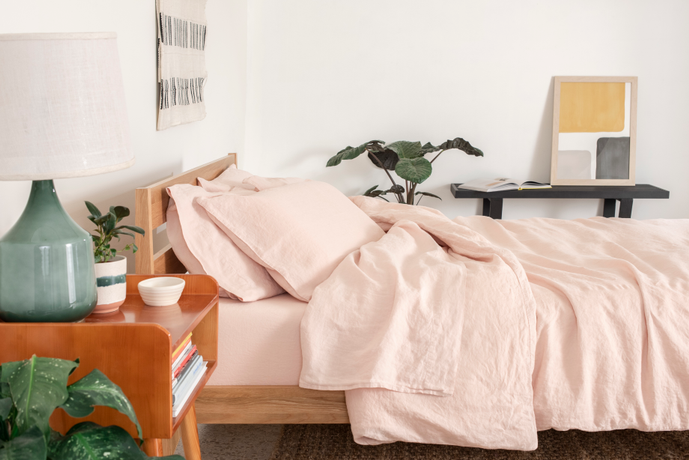 Side view of a Tuft & Needle bed and nightstand. The bed is made with pink bedding and the nightstand has a green based lamp with a white shade sitting on top of it.