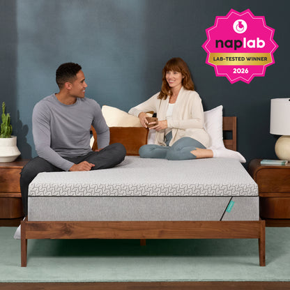 Two people sitting on a Tuft and Needle bed with a Naplab award badge, against a gray wall.