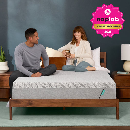 Two people sitting on a Tuft and Needle bed with a Naplab award badge, against a gray wall.