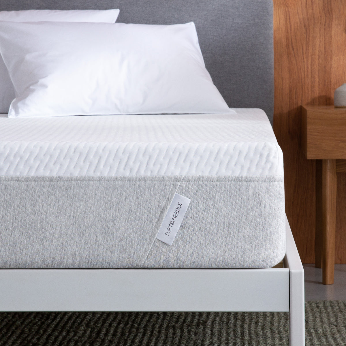 Close up of a Tuft and Needle  mattress on a white bed frame and grey headboard with two pillows on top of it.
