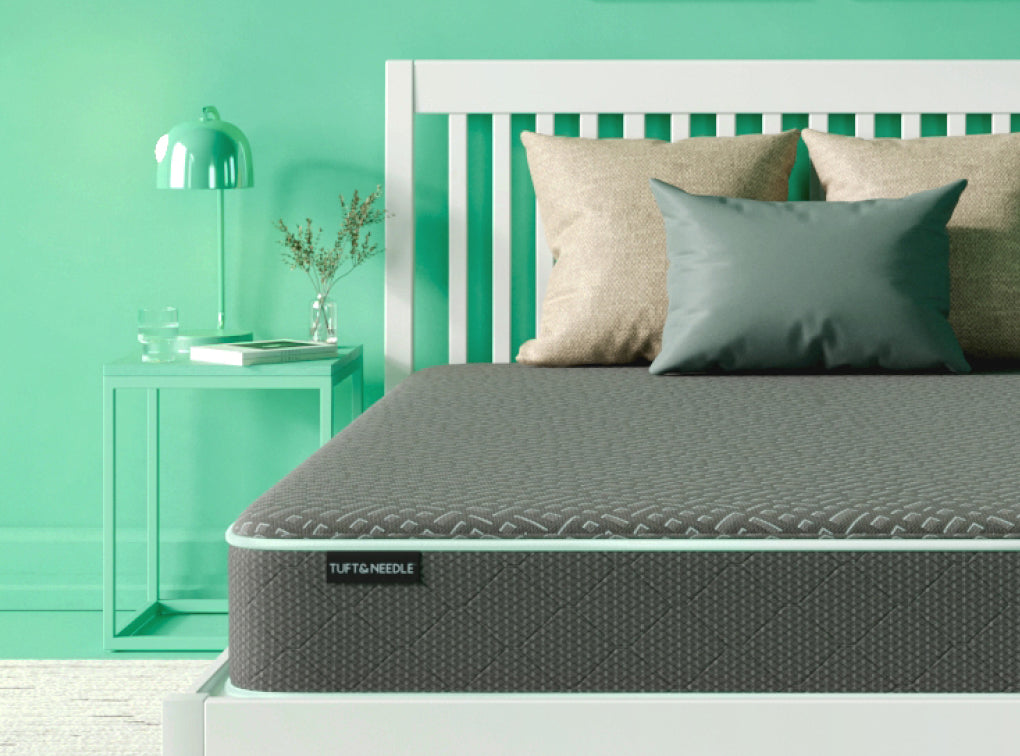 Tuft and Needle Essential mattress sitting on a white wood bedframe with 3 pillows on top of it.