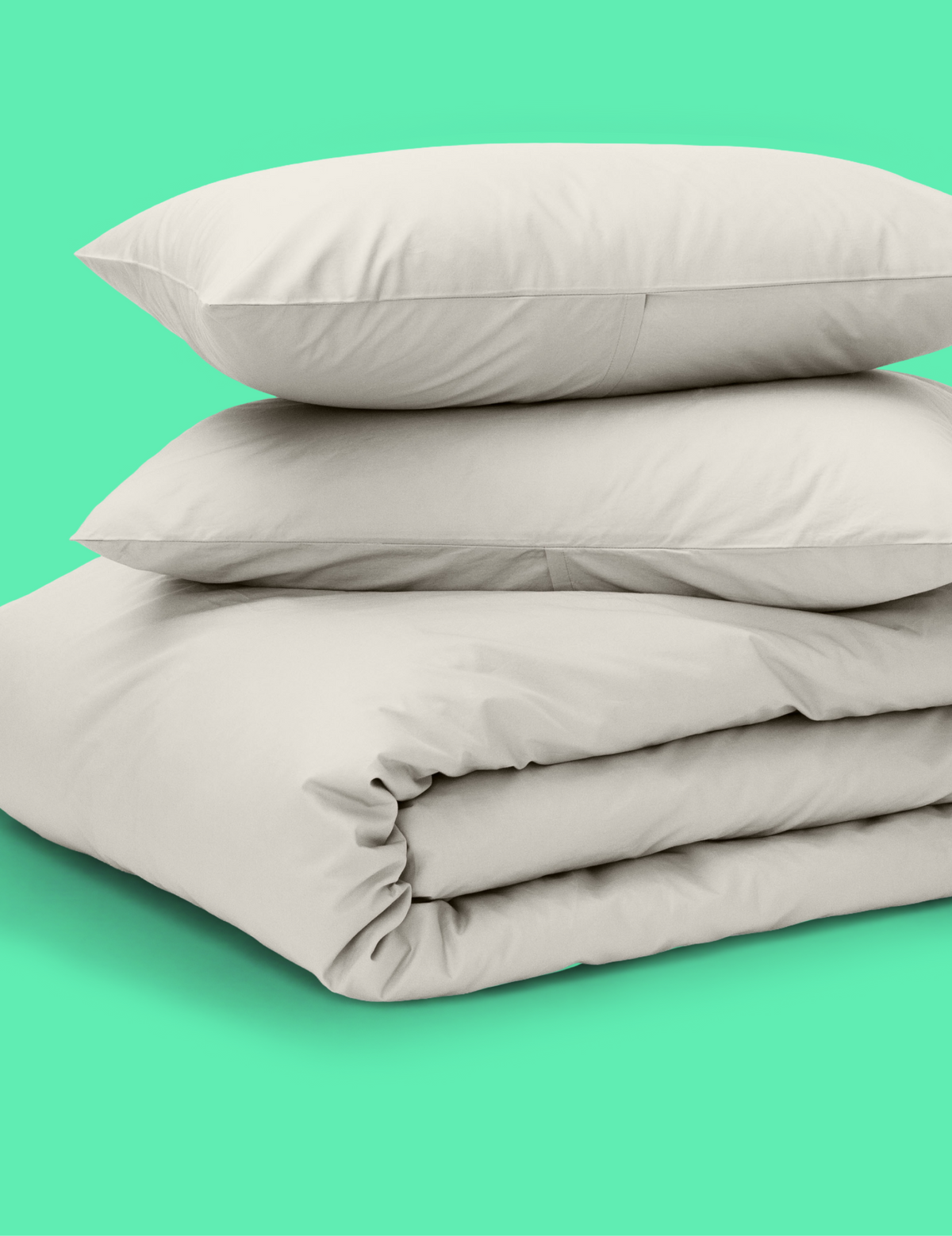 A stack of light beige Tuft and Needle bedding, including two pillows and a folded duvet cover, set against a solid mint green background.