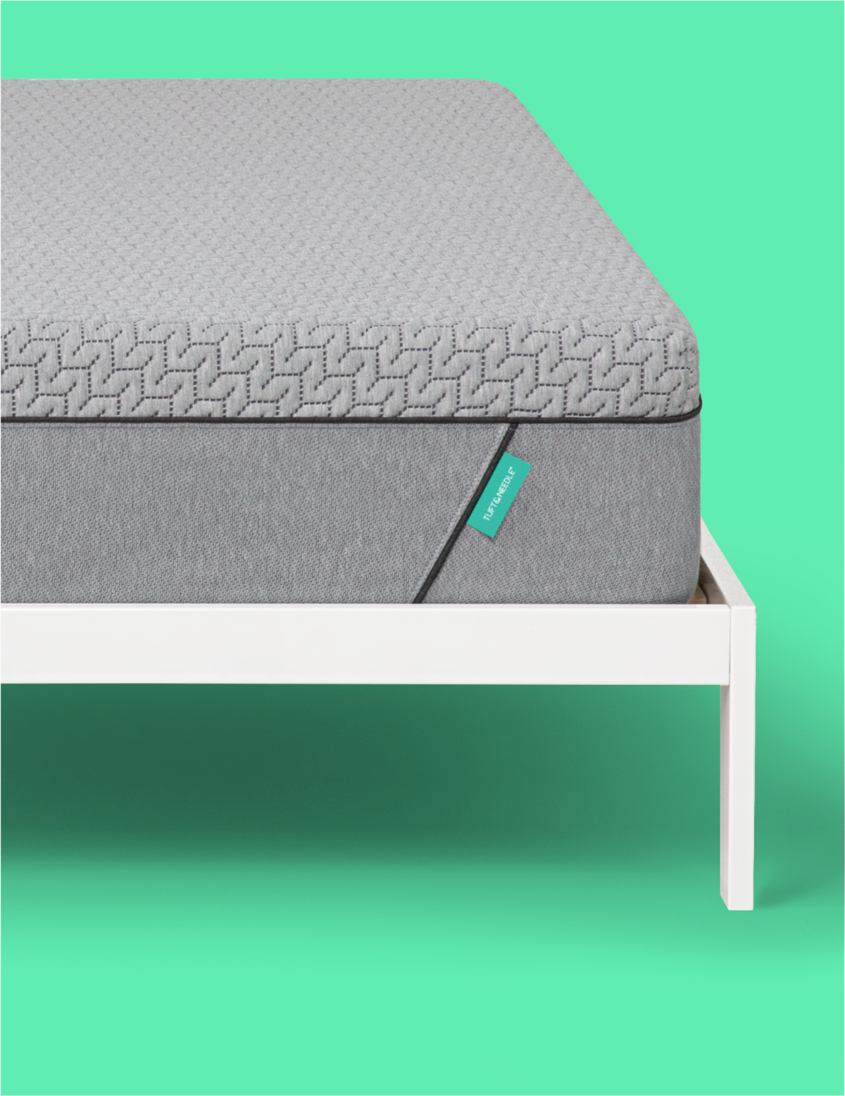 A close-up of a gray Tuft and Needle mattress on a white bed frame against a solid mint green background, with a small teal tag on the side of the mattress.
